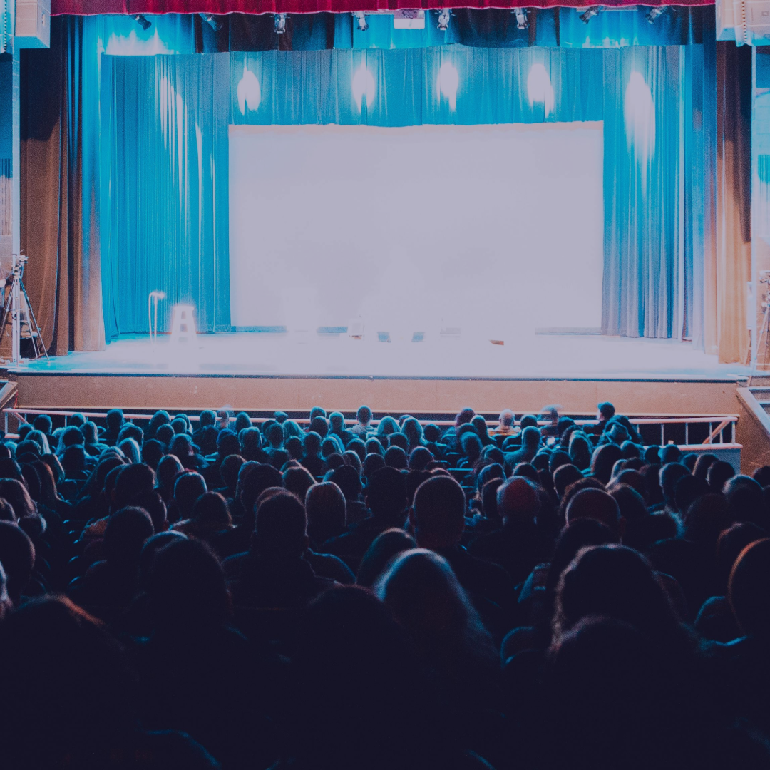 Theater audience viewed from the rear stalls, stage lit in blue, full house